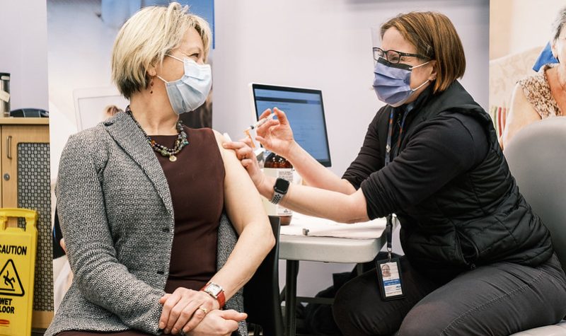 A woman wearing black gives a vaccine shot to Dr. Bonnie Henry in her arm in a medical clinic