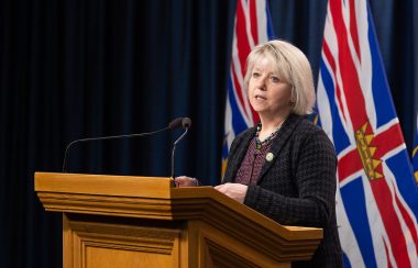 Photo of Dr. Bonnie Henry giving a press conference to the media standing in front of two British Columbian flags.