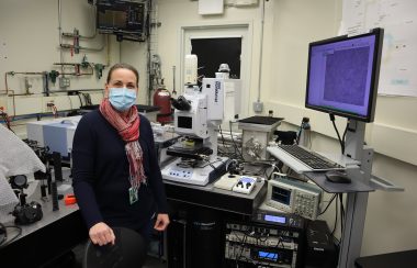 A woman is standing infront of a medical piece of machinery called an MRI - light beam which calculates brain health