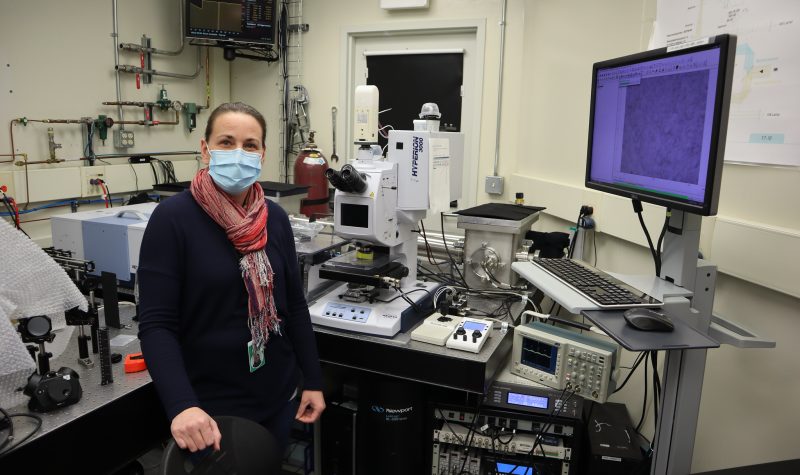 A woman is standing infront of a medical piece of machinery called an MRI - light beam which calculates brain health