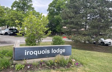Street sign of Iroquois Lodge, flowers and grass surround horizontal grey sign with white lettering. Trees across the entrance way in background hide the building.