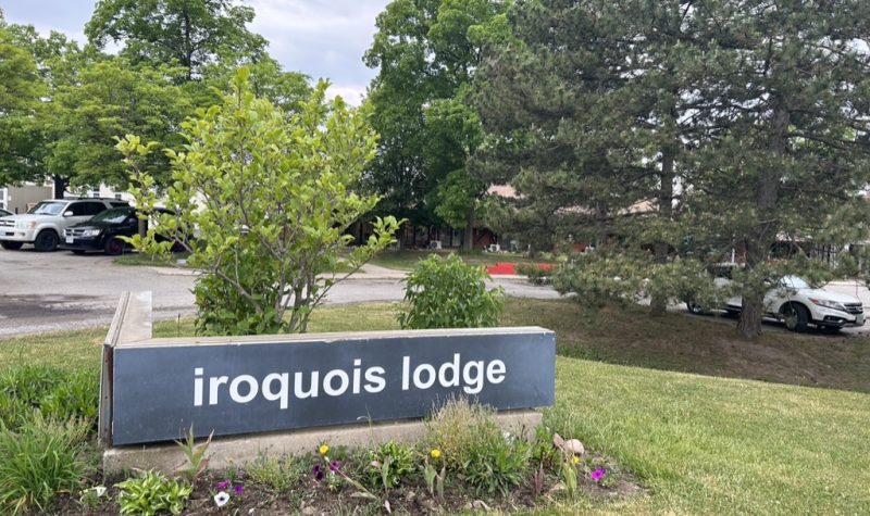Street sign of Iroquois Lodge, flowers and grass surround horizontal grey sign with white lettering. Trees across the entrance way in background hide the building.