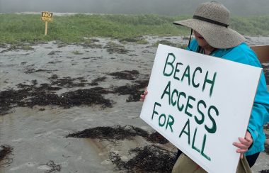 A woman stands on a beach holding a sign demanding beach access for all