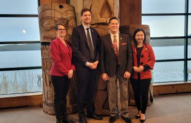 4 people stand in front of traditional indigenous carvings