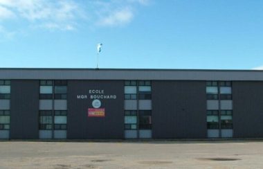 École primaire Mgr Bouchard, bâtisse grise foncée dans un stationnement vide avec un ciel bleu dégagé en arrière plan.