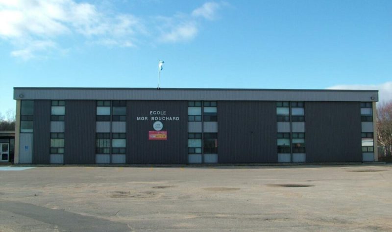 École primaire Mgr Bouchard, bâtisse grise foncée dans un stationnement vide avec un ciel bleu dégagé en arrière plan.