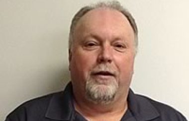 A headshot of Ed Walsh standing in front of a bare wall and wearing a grey golf shirt.