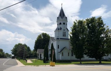 On peut voir l'église de bois St. Patrick's de Perkinsfield. L'église est peinte en blanc avec le toit noir et est de style néo-gothique. On voit les arbres et le gazon du terrain de l'église à proximité ainsi que les route de comté 6 et 10.