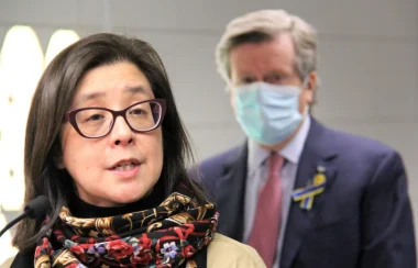 A person with a colourful scarf and glass rim glasses speaks with a person behind them on the right wearing a suit and a blue mask.