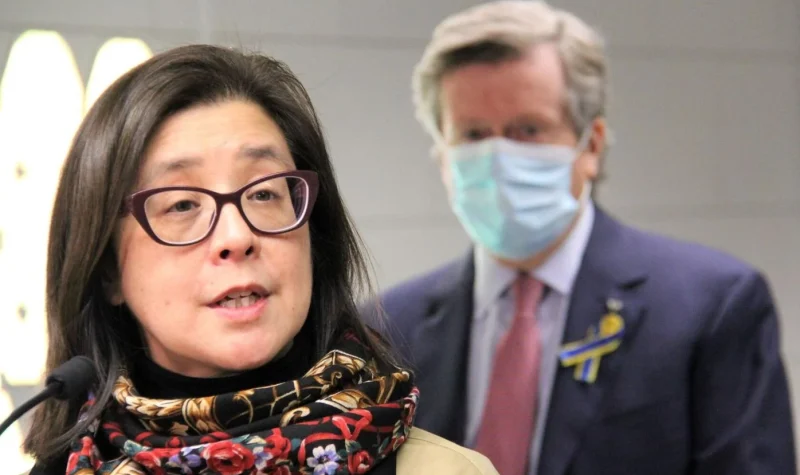A person with a colourful scarf and glass rim glasses speaks with a person behind them on the right wearing a suit and a blue mask.