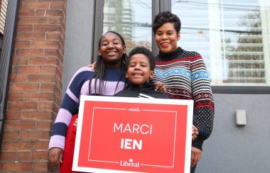 Three people outside a house holding a red sign