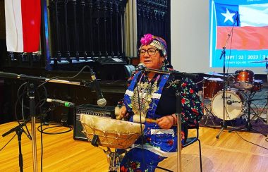 mujer mapuche cantando y tocando un tambor frente a unas banderas chilenas