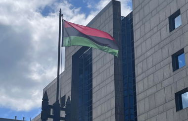 A flag waving in the wind with the blue sky a large building in the background.