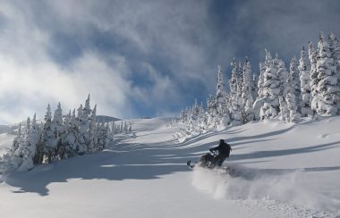 A blue sky is nearly hidden by whispy clouds on a bright day. A snowmobiler is charging through fresh powder on a mountain in the Bulkley Valley.