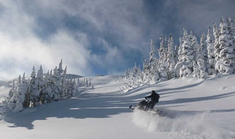 A blue sky is nearly hidden by whispy clouds on a bright day. A snowmobiler is charging through fresh powder on a mountain in the Bulkley Valley.