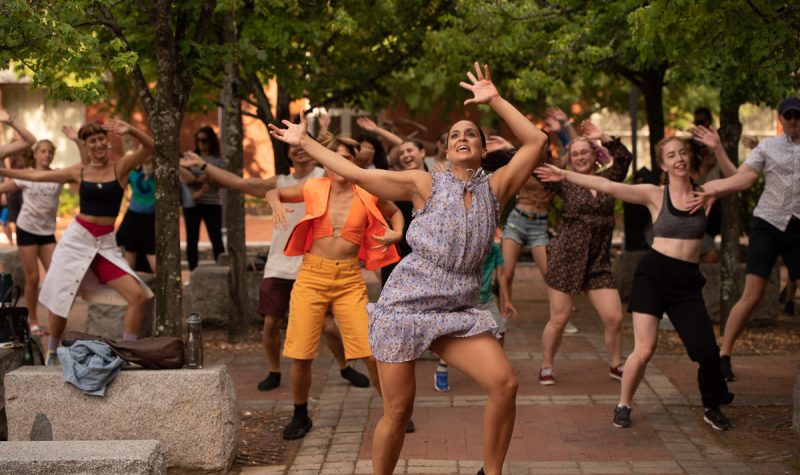 Une dame avec robe picotter de couleur taupe dansant les bras en l'air au centre de la photo. En arrière d'elle, plusieurs personnes font la même danse. Tout cela dans le centre d'arbres sur trottoir en brique et ciment.