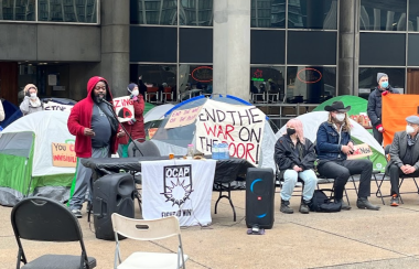 People sit in front of pitched tents of various colours with signs and a building in the background.