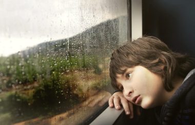 Un garçon est appuyé contre une fenêtre sur laquelle on voit des gouttes de pluie. Il regarde à l’extérieur.