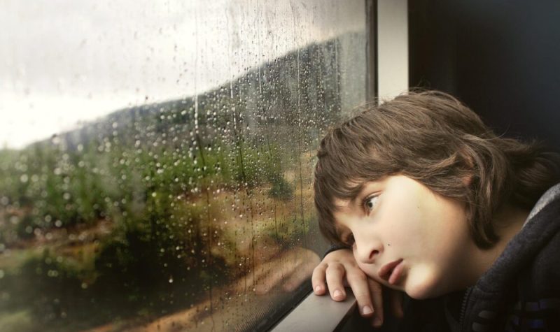 Un garçon est appuyé contre une fenêtre sur laquelle on voit des gouttes de pluie. Il regarde à l’extérieur.