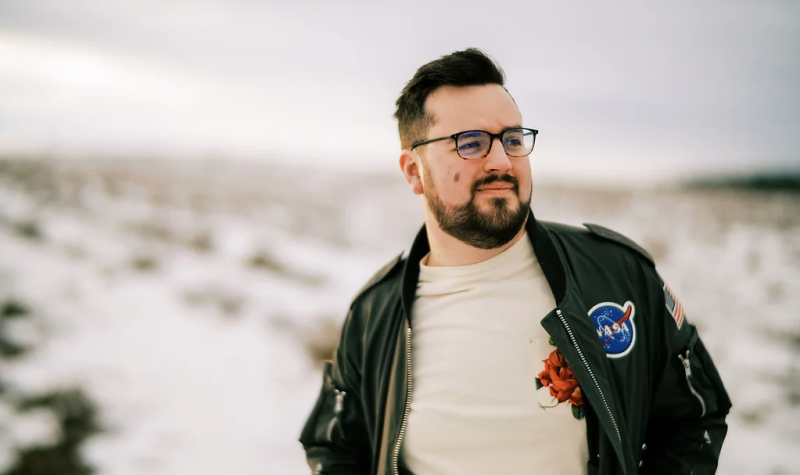 A man in a white tshirt and black jacket stands before a blurred out backgorund of an overcast day, there is snow on the ground. He Wears a white T-shirt and a black leather jacket with a NASA patch over the left breast. He looks to his left as he wears black rimmed glasses and a dark short beard.