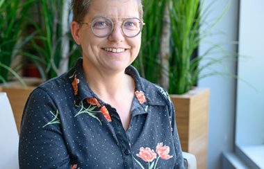 Pictured is Dr. Estelle Chamoux sitting on a chair with tall green plants serving as the background.