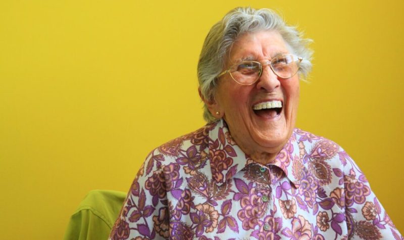 Une personne âgée rit aux éclats devant un mur jaune.