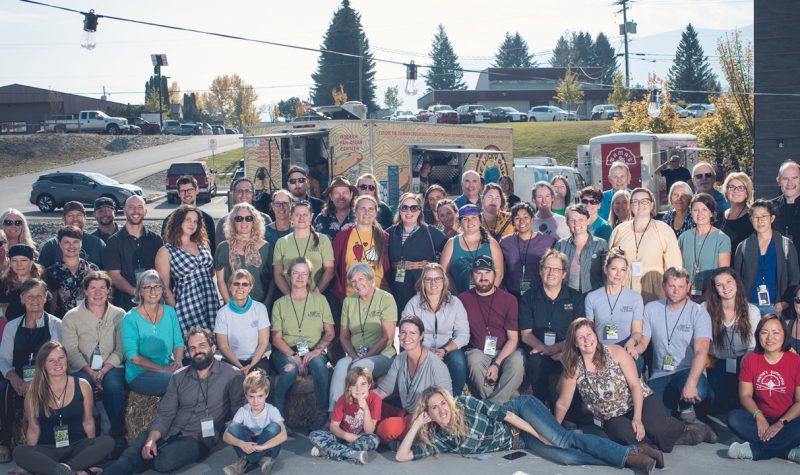Group of people gathered on a city street for a photo. Food trucks, trees and a distant mountain is in the background.