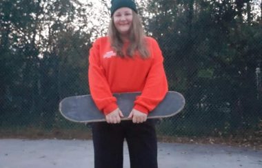 A woman wearing a red shirt and toque holding a skateboard and smiling.