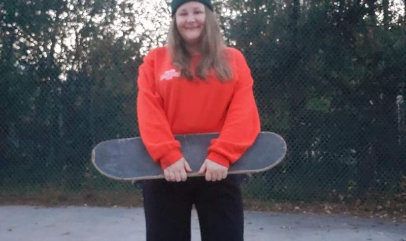 A woman wearing a red shirt and toque holding a skateboard and smiling.