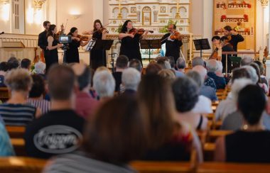 Au premier plan, le public observant les musiciens jouer sur scène, en intérieur, situés au premier plan