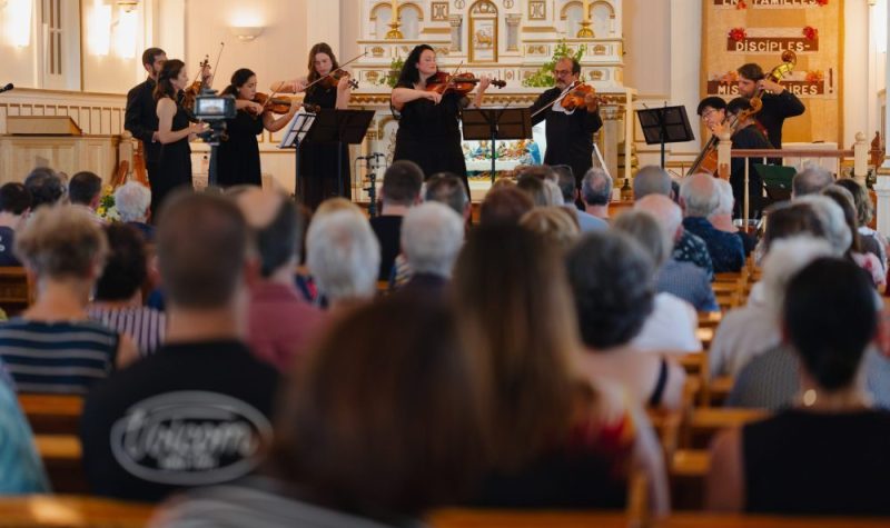 Au premier plan, le public observant les musiciens jouer sur scène, en intérieur, situés au premier plan