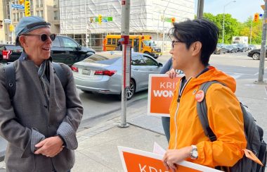 A person on the right with an orange jacket and sign speaking to a person on the left wearing grey and glasses.