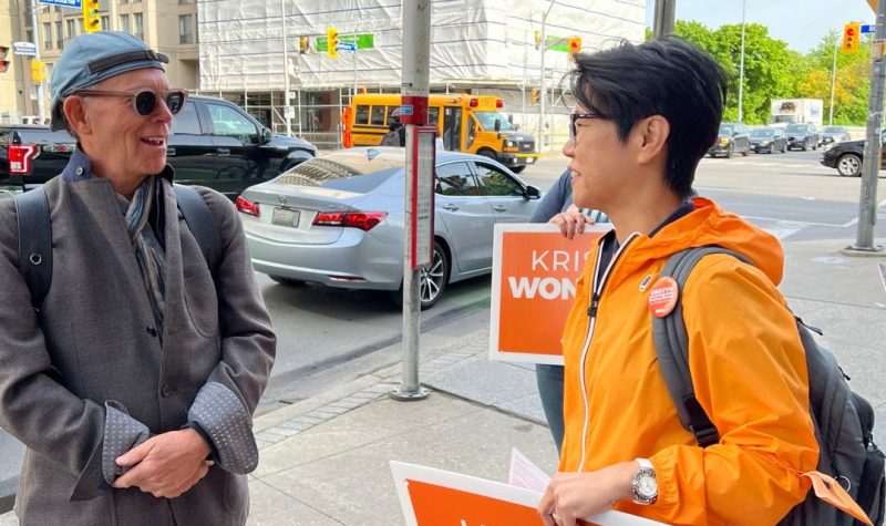 A person on the right with an orange jacket and sign speaking to a person on the left wearing grey and glasses.