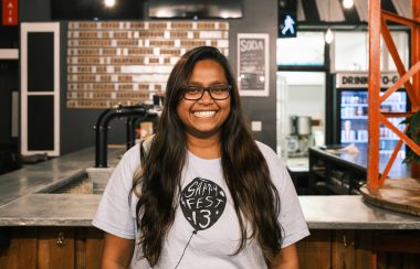 A woman with a big smile wearing a Sappyfest 13 t-shirt stands in front of a bar.