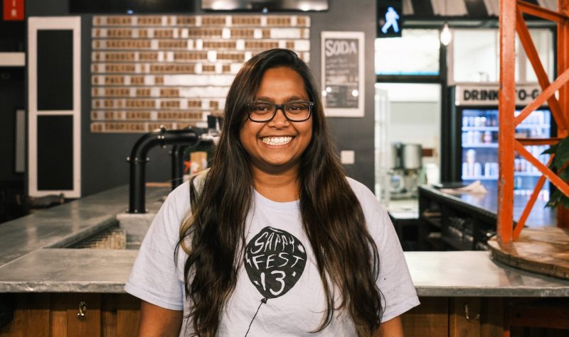 A woman with a big smile wearing a Sappyfest 13 t-shirt stands in front of a bar.