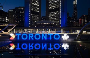 A blue sign of letters with a white leaf against a backdrop of buildings at night.