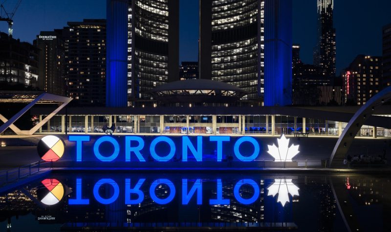 A blue sign of letters with a white leaf against a backdrop of buildings at night.