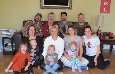 Madame Carole Desgagnésau centre accompagnée des 11 autres membres de sa famille. Ils sont assis sur un divan brun et un plancher de bois. Il y a un mur jaune et un cadre noir derrière.