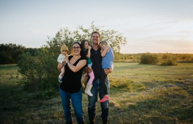 La famille enlacée dans un champ en été, les parents tiennent les trois enfants dans leurs bras.