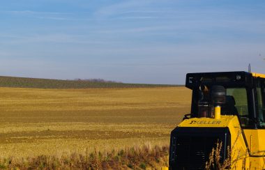 A dusty brown barren field with vegetation coverage further behind it. The sky is blue with wisps of white clouds. Half of a yellow and black painted canopied tractor sits in the foreground amongst dry plant stalks.