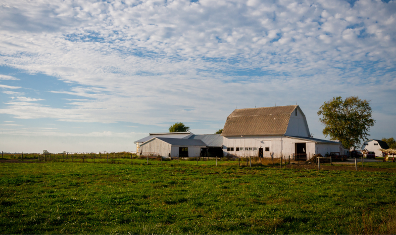 A white barn sits on farmland underneath a partly cloudy blue sky.