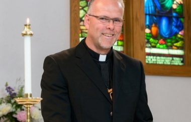 Allan Doerken stands in a church next to a candle and stained glass windows.