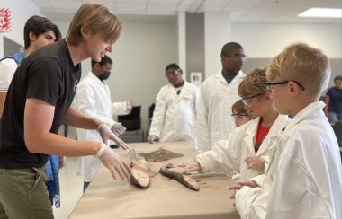 A group photo of 8 people, 6 children are wearing lab coats and touching sharks. A marine biologist is explaining sharks and has his hands on the table. This is in a classroom with many chairs and tables.