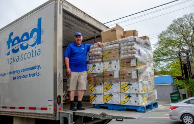 A man dressed in Feed Nova Scotia shirt and cap, standing inside a Feed Nova Scotia white truck with packages of food reaching over his head. There are about 30 crates of eggs and produce and other products.
