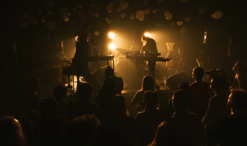 Dans un décor à l'éclairage tamisé, une foule assiste à un spectacle. Sur la scène, deux musiciens s'y trouvent avec une batterie et un clavier.
