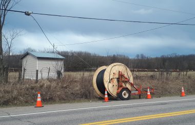 A large spool of cable sits by the side of the road, with four pylons marking it, and the end of the spool connected to nearby telephone wires.