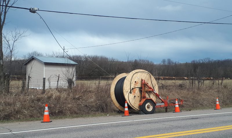 A large spool of cable sits by the side of the road, with four pylons marking it, and the end of the spool connected to nearby telephone wires.