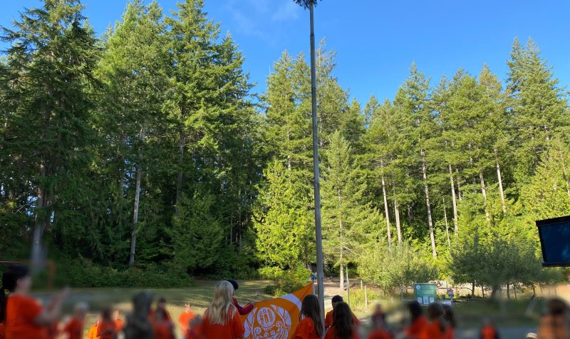 Several people with orange shirts surround a Truth and Reconciliation Flag as it is being raised on a flagpole.