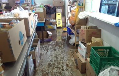 Cardboard boxes of food sit on stainless steel counters and shelves. The floor is covered in suds and water.