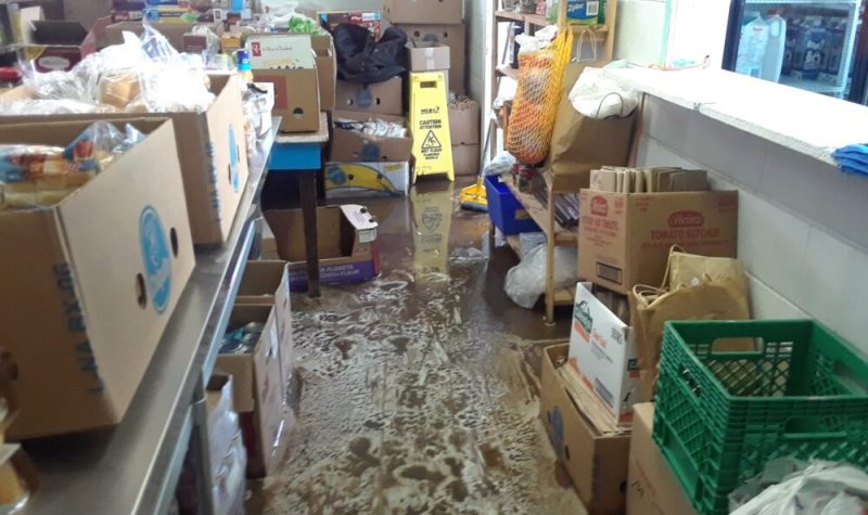 Cardboard boxes of food sit on stainless steel counters and shelves. The floor is covered in suds and water.